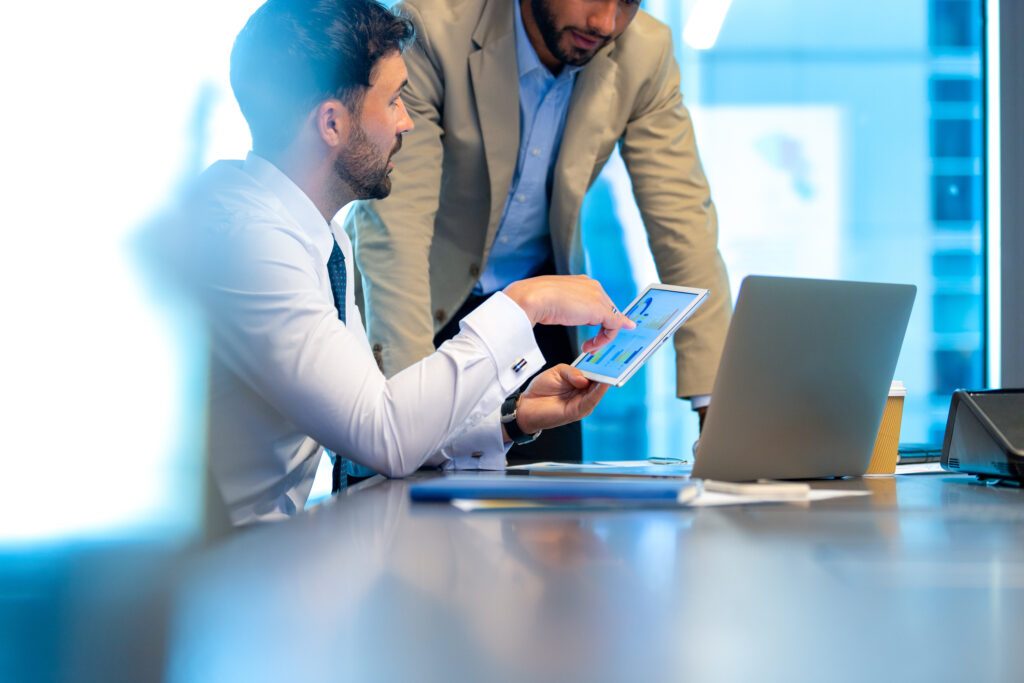 2 business men working in the office. There is a laptop on the table and one businessman is holding a digital tablet with finance charts and graphs